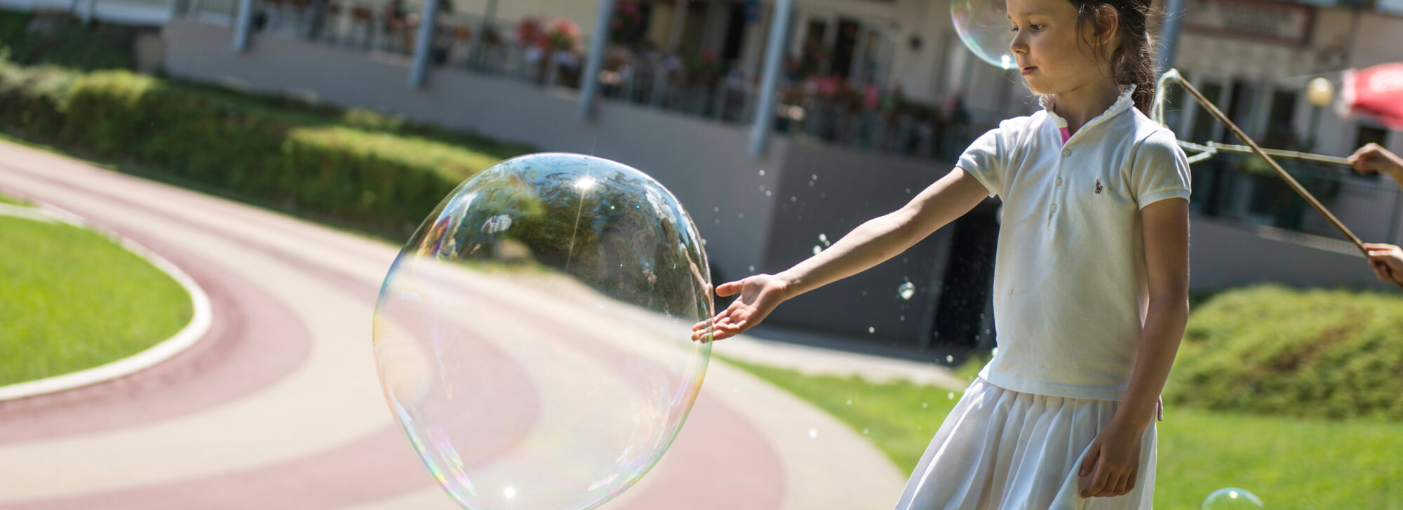 Ein Kind spielt draußen bei Sonnenschein mit einer großen, durchsichtigen Seifenblase, die reflektiert. Der Hintergrund zeigt einen geschwungenen Weg und Grünflächen.