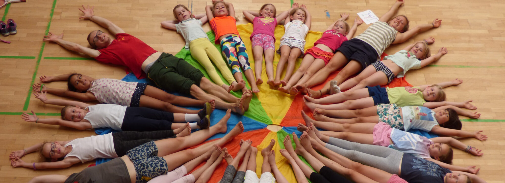 Eine fröhliche Gruppe von Kindern und einem Erwachsenen liegt bei einem Kindergeburtstag im Kreis auf dem Boden. In der Mitte liegt ein buntes Fallschirmtuch als Teil eines Gemeinschaftsspiels.