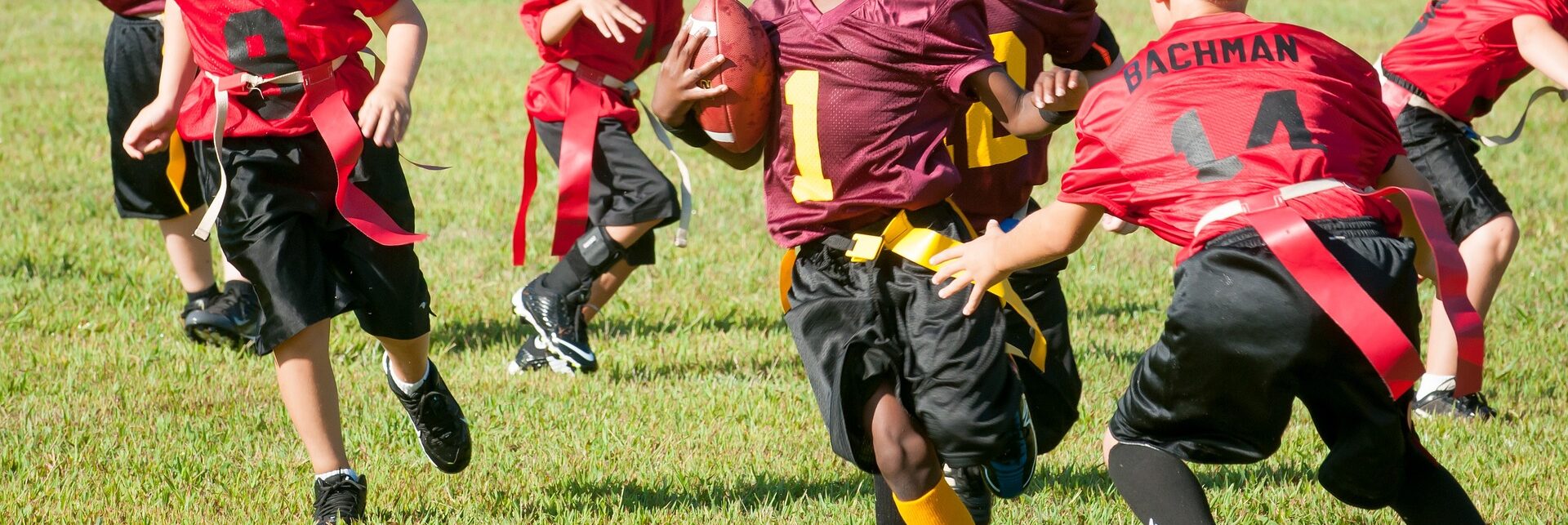 Kinder spielen Flag Football auf einem Rasenfeld. Ein Spieler in einem burgunderfarbenen Trikot trägt den Ball, während andere Kinder in roten Trikots versuchen, seine Flagge zu greifen.