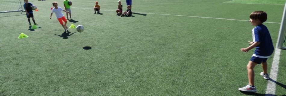 Kinder spielen Fußball auf einem Kunstrasenplatz unter freiem Himmel bei sonnigem Wetter.