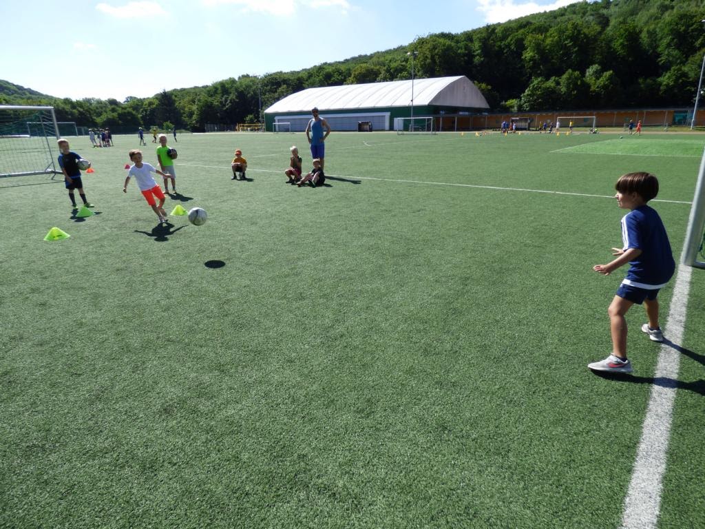 Kinder spielen Fußball auf einem Kunstrasenplatz unter freiem Himmel bei sonnigem Wetter.