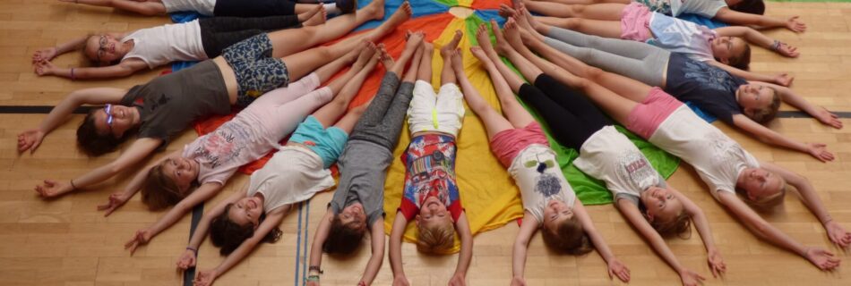 Eine Gruppe von Kindern und Jugendlichen liegt kreisförmig auf dem Boden einer Turnhalle, mit den Füßen in der Mitte und den Armen ausgestreckt. Sie liegen auf einem bunten Schwungtuch, das sich sternförmig unter ihnen ausbreitet.