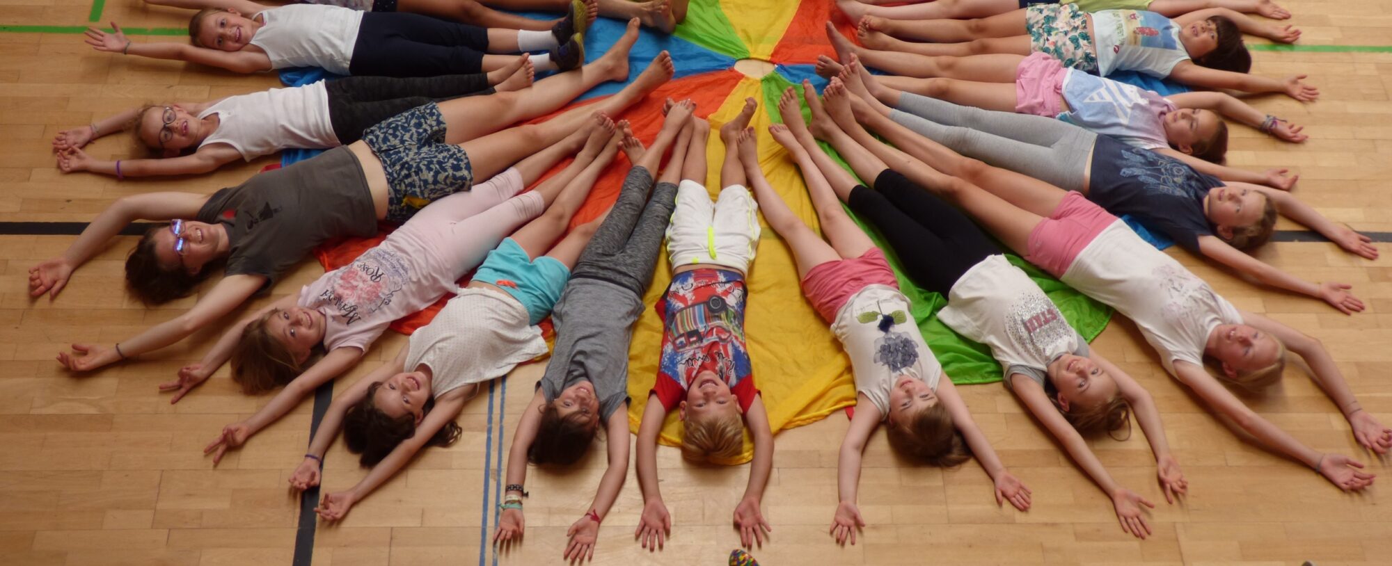 Eine Gruppe von Kindern und Jugendlichen liegt kreisförmig auf dem Boden einer Turnhalle, mit den Füßen in der Mitte und den Armen ausgestreckt. Sie liegen auf einem bunten Schwungtuch, das sich sternförmig unter ihnen ausbreitet.