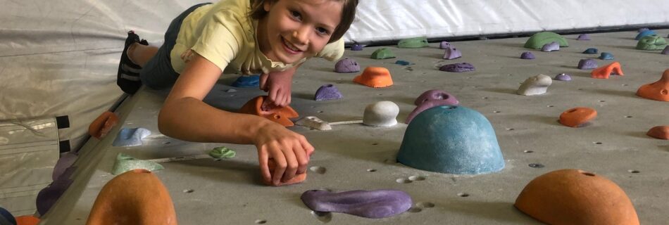 Ein junges Mädchen liegt auf einer Boulderwand und greift nach einem Griff, während sie in die Kamera lächelt. Die Wand ist mit bunten, unterschiedlich geformten Klettergriffen versehen.
