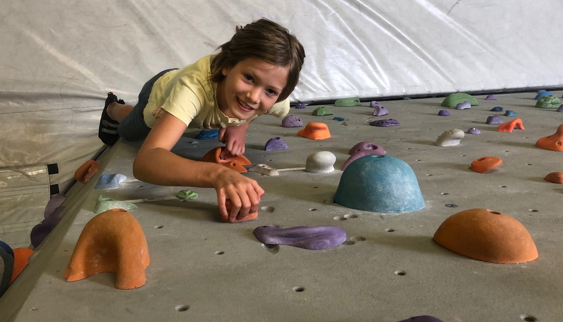 Ein junges Mädchen liegt auf einer Boulderwand und greift nach einem Griff, während sie in die Kamera lächelt. Die Wand ist mit bunten, unterschiedlich geformten Klettergriffen versehen.