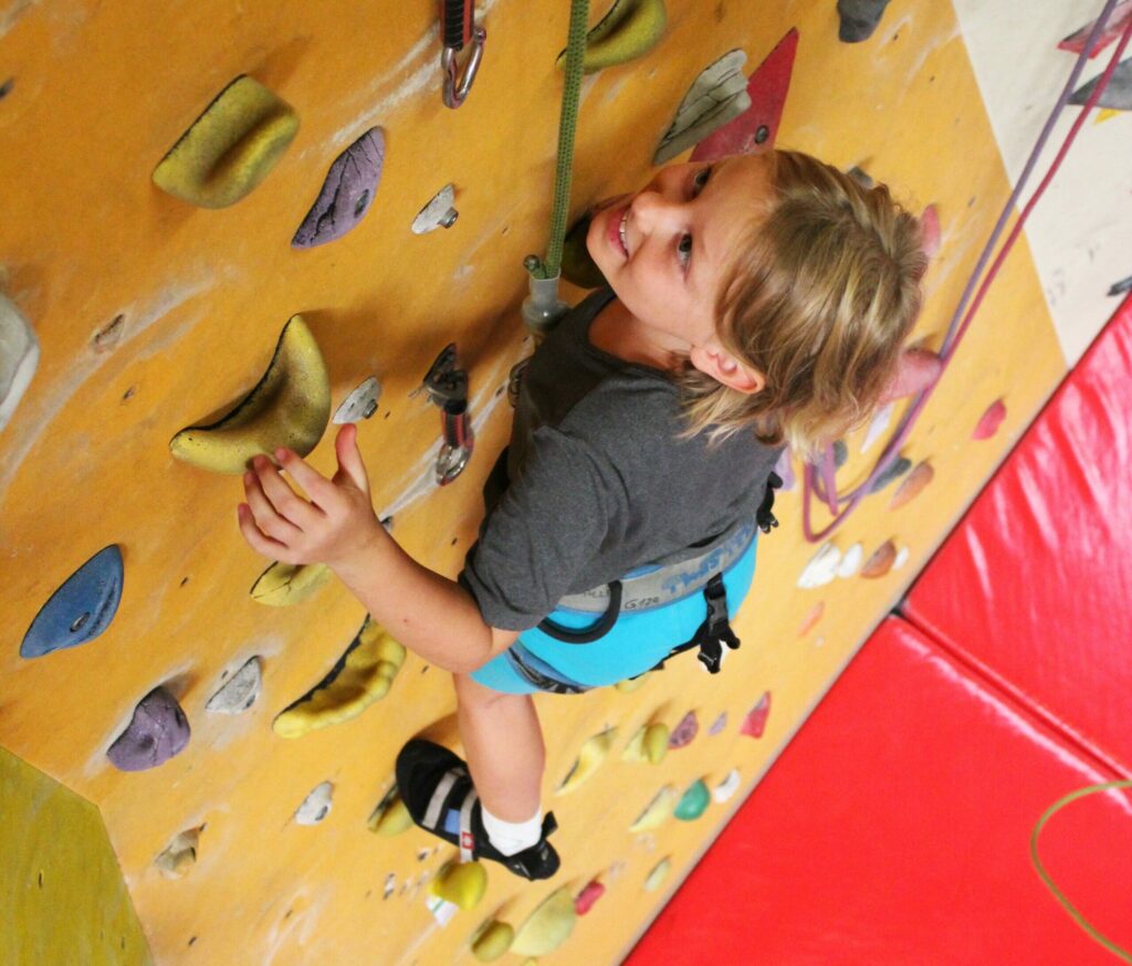 Ein Mädchen klettert mit einem Klettergurt gesichert an einer bunten Indoor-Kletterwand.