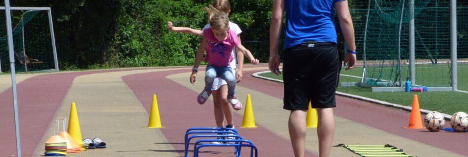 Ein Mädchen springt über Mini-Hürden auf einer Tartanbahn, während ein Trainer in Sportkleidung barfuß daneben steht und zuschaut.