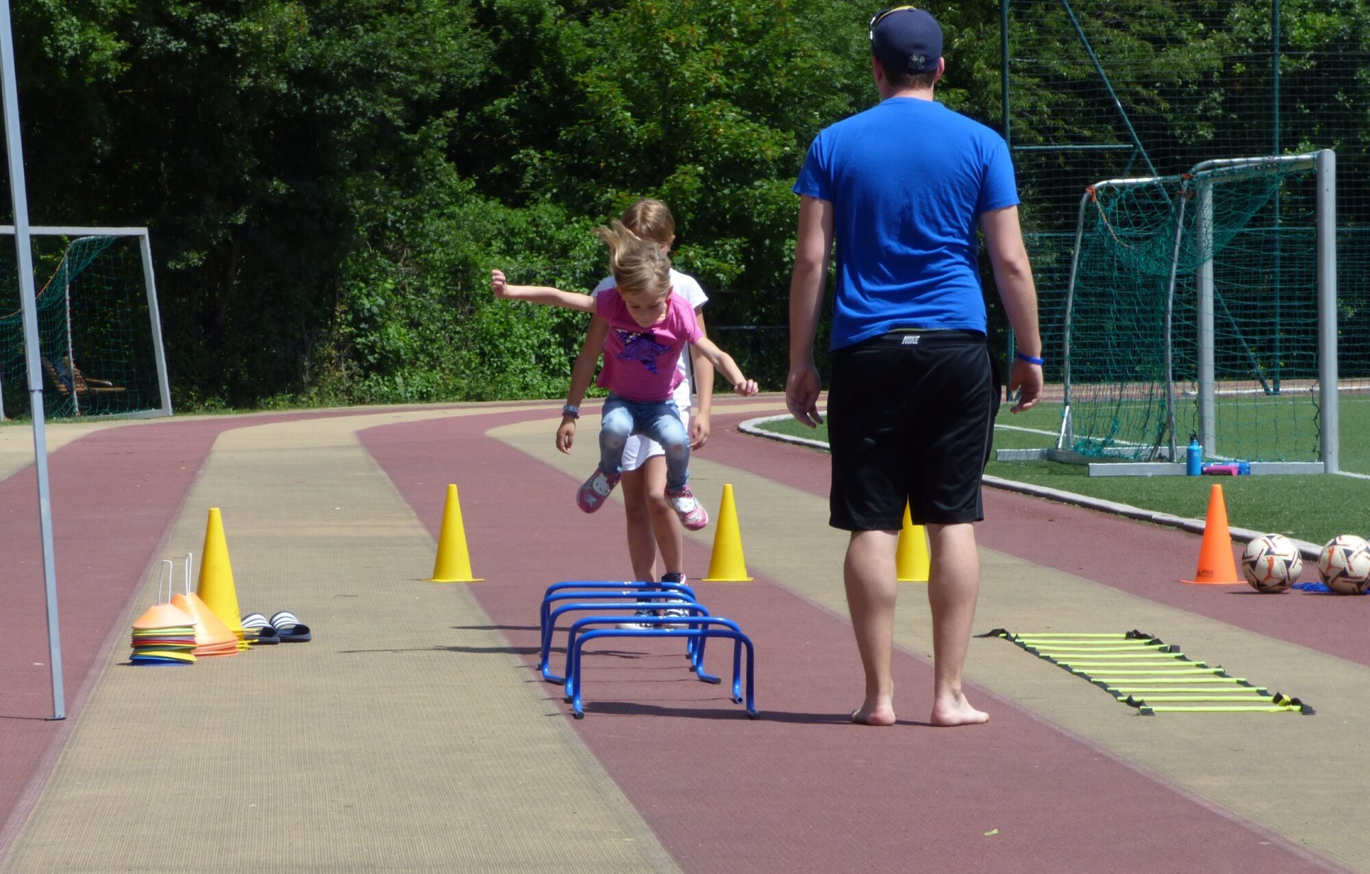 Ein Mädchen springt über Mini-Hürden auf einer Tartanbahn, während ein Trainer in Sportkleidung barfuß daneben steht und zuschaut.