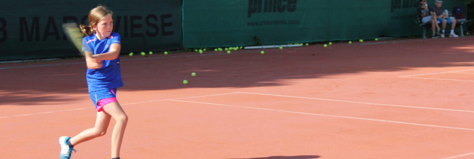 Ein Mädchen in blauer Sportkleidung schlägt auf einem roten Tennisplatz mit einem Schläger einen Ball, während im Hintergrund mehrere Tennisbälle und Zuschauer zu sehen sind.