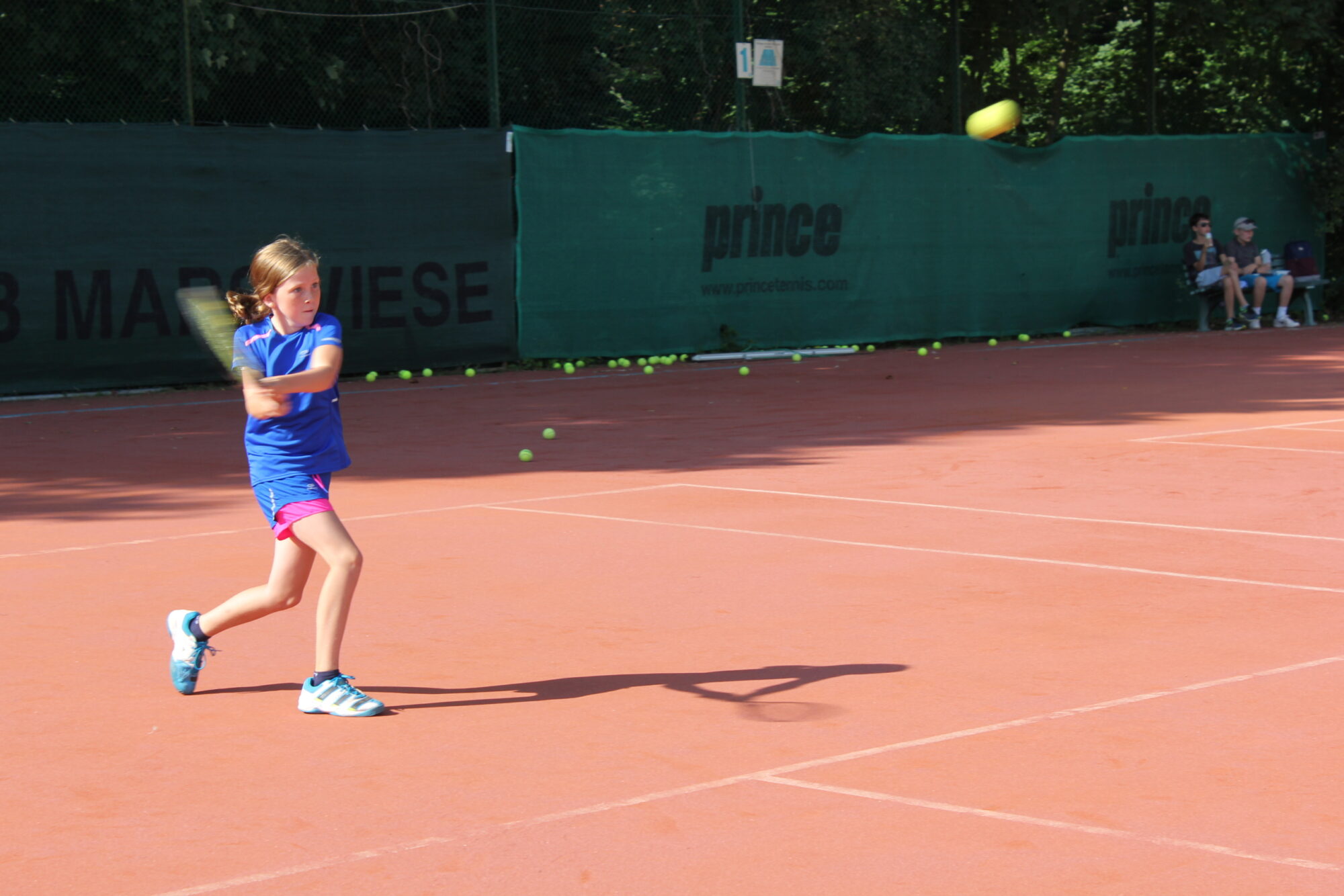 Ein Mädchen in blauer Sportkleidung schlägt auf einem roten Tennisplatz mit einem Schläger einen Ball, während im Hintergrund mehrere Tennisbälle und Zuschauer zu sehen sind.