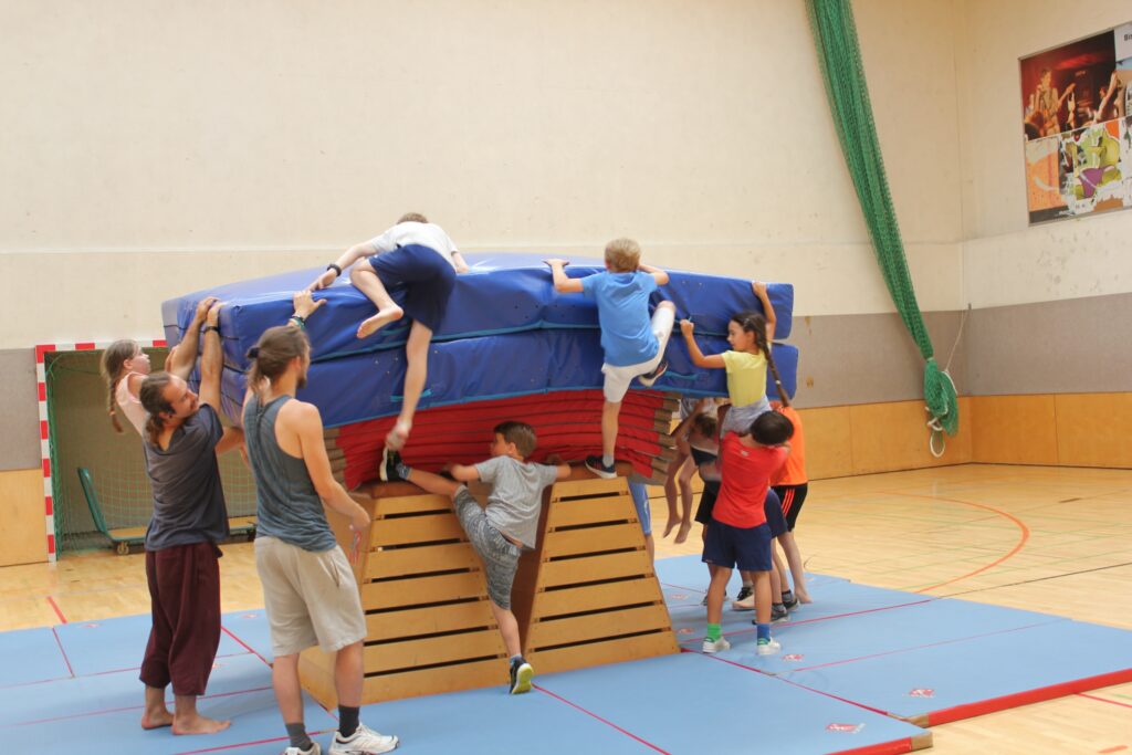 Kinder und Betreuer in einer Turnhalle bauen eine Kletter- und Sprungstation aus Weichbodenmatten und einem Holzkasten.
