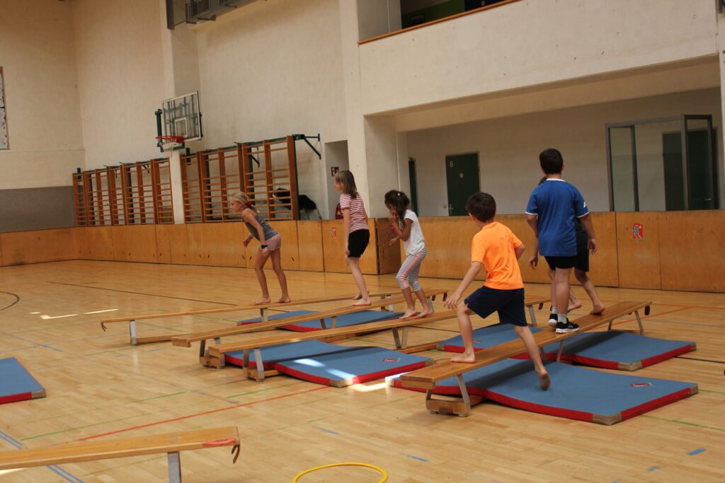 Kinder balancieren barfuß auf Turnbänken, die über Weichbodenmatten gelegt sind, in einer Turnhalle.