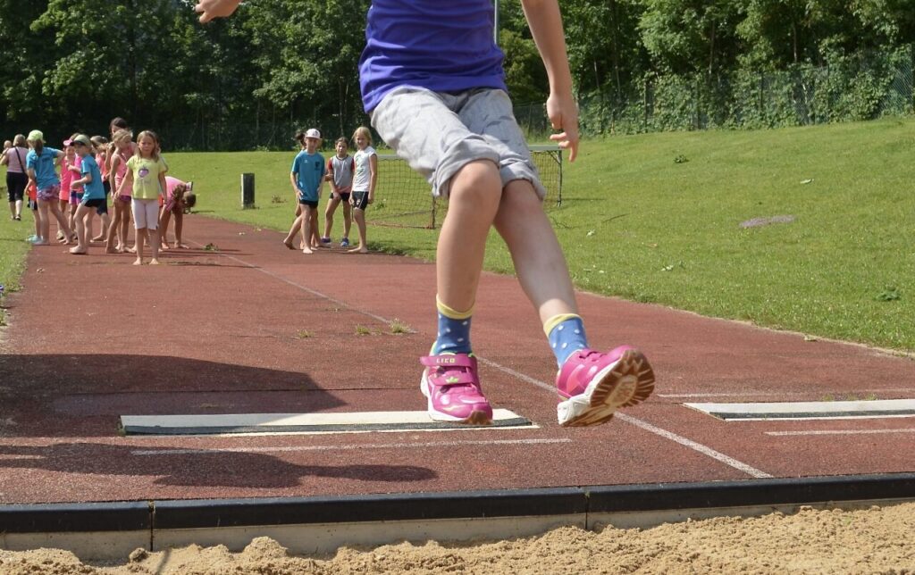 Ein Kind springt in eine Weitsprunggrube, während andere Kinder im Hintergrund anstehen und zuschauen.
