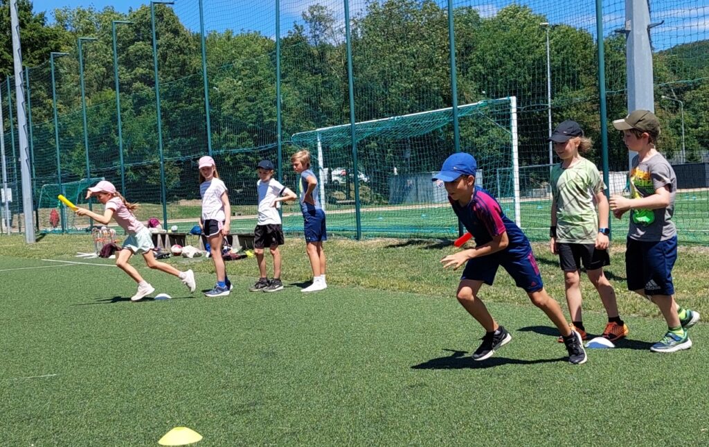 Kinder stehen in zwei Reihen auf einem Sportplatz und starten ein Staffellauf-Rennen mit bunten Staffelstäben in der Hand.