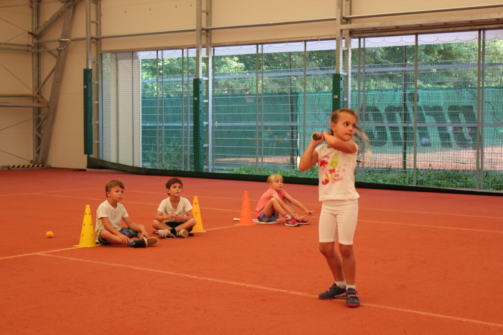 Ein Mädchen steht in Schlagposition mit einem Tennisschläger auf einem Hallenplatz, während drei andere Kinder auf dem Boden sitzen und zusehen. Um sie herum sind bunte Hütchen aufgestellt.