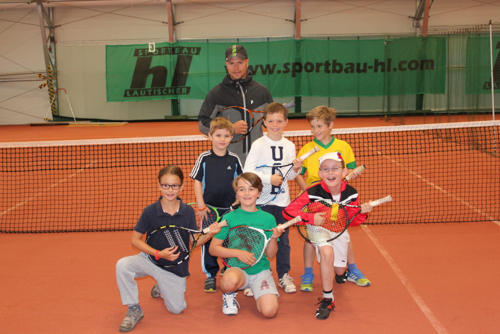 Eine Gruppe von sechs Jungen mit Tennisschlägern posiert gemeinsam mit einem Trainer in einer Tennishalle vor einem Netz.